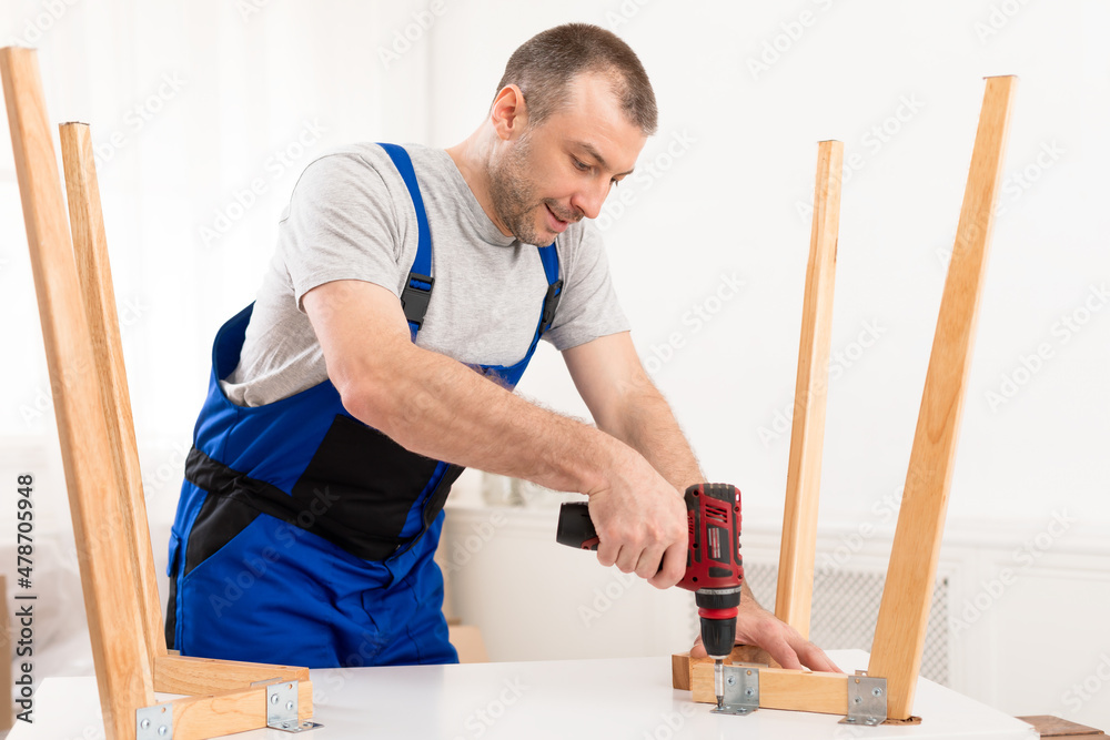 Repairman In Blue Uniform Fixing Table With Electric Drill Indoors Repairman In Blue Uniform Fixing Table With Electric Drill Indoors