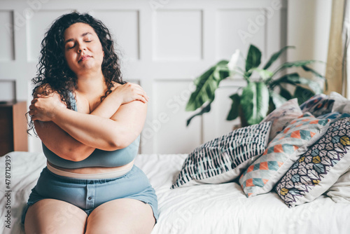 Curly haired overweight young woman in grey top and shorts with satisfaction on face accepts curvy body shape standing in stylish bedroom closeup