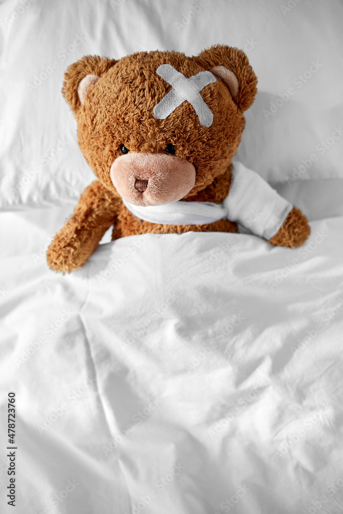 medicine, healthcare and childhood concept - ill teddy bear toy with bandaged paw and medical patch on head lying in bed