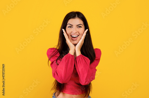 Wow offer. Amazed armenian woman with open mouth touching her cheeks with hands over yellow background
