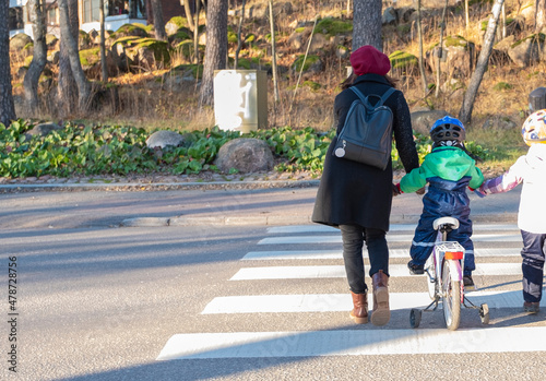 A young woman with children, a boy and a girl, cross the city road along a pedestrian crossing. Children ride a bike.