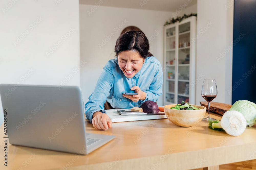 Woman holding smart phone laughing on video call using laptop at home Woman holding smart phone laughing on video call using laptop at home