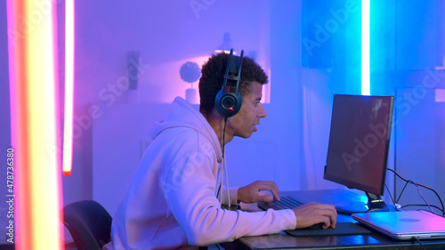 Profile view of young gammer in headphones playing in online shooter. The room have warm colored neon light. photo