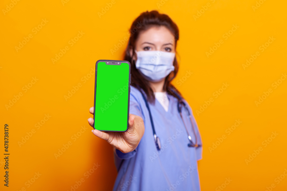 Nurse holding vertical green screen on smartphone while wearing face mask against coronavirus. Medical assistant showing device with chroma key and isolated mockup template on camera. Nurse holding vertical green screen on smartphone while wearing face mask against coronavirus. Medical assistant showing device with chroma key and isolated mockup template on camera.