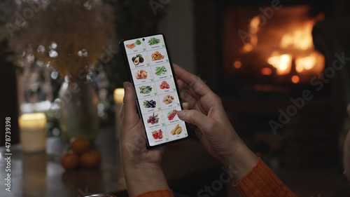 Ordering food using a smartphone at home. A woman selects vegetables in an online store using an application on a smartphone. Home evening furnishings with a burning fire in the fireplace.