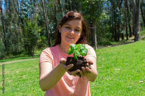 Beautiful young Hispanic woman holding a small plant in her field hands before being planted in a green field surrounded by trees
