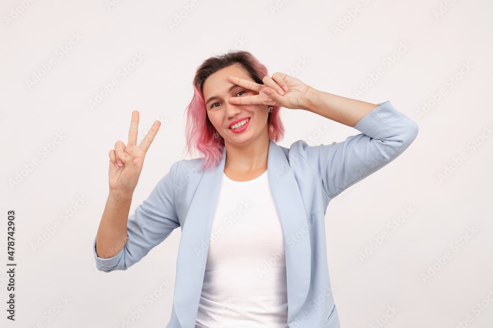 Fototapeta premium Successful businesswoman showing peace, v-sign salute near eye and smiling confident, standing in suit against white background