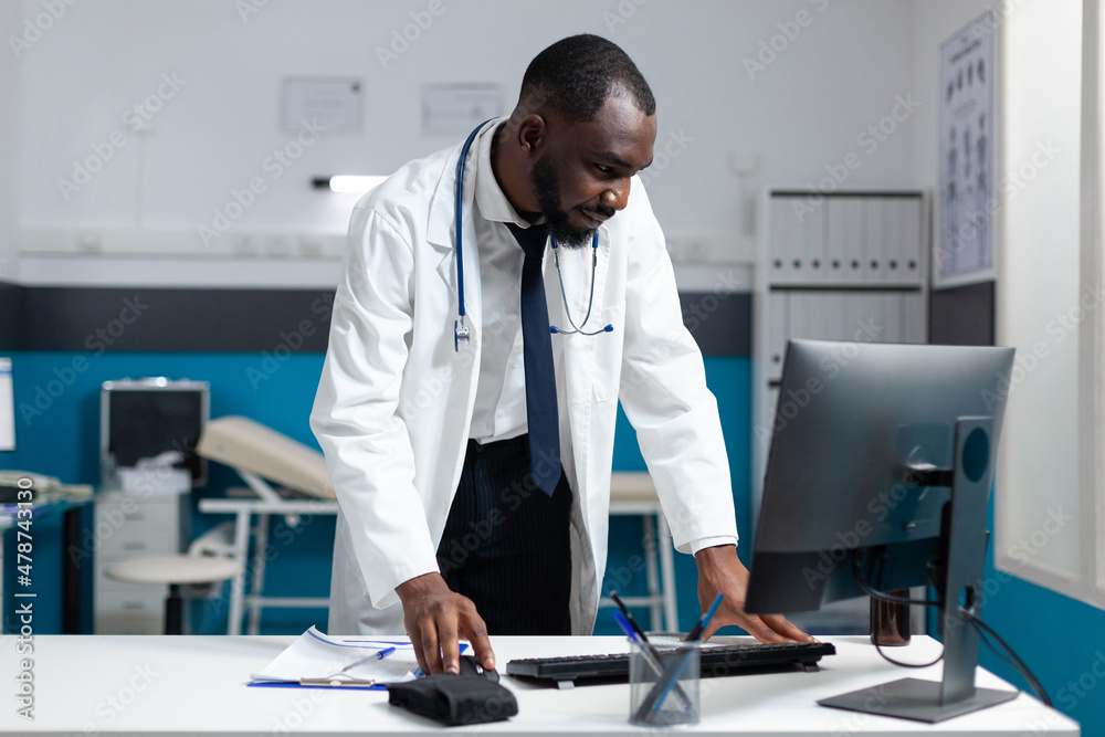 African american practitioner man monitoring disease report on computer analyzing healthcare treatment during clinical appointment. Doctor working at medical expertise in hospital office African american practitioner man monitoring disease report on computer analyzing healthcare treatment during clinical appointment. Doctor working at medical expertise in hospital office