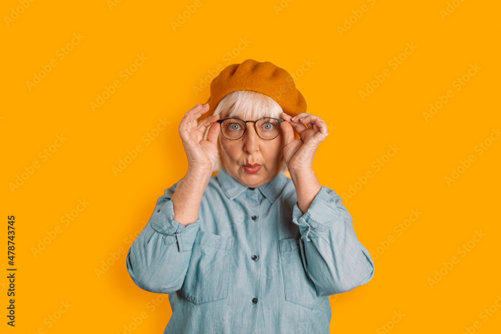 Photo of blonde hair 50s woman astonished face touch glasses look camera isolated over orange color background Photo of blonde hair 50s woman astonished face touch glasses look camera isolated over orange color background