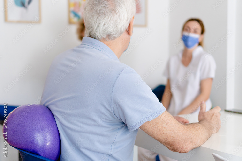 Obraz premium older man sitting with a pilates and yoga medicine ball on his back to rehabilitate from shoulder tendonitis, while talking to the occupational therapist, a physiotherapist