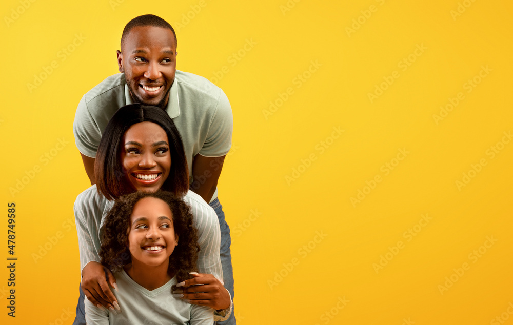 Positive african american family of three looking aside at empty space on yellow background, standing behind each other