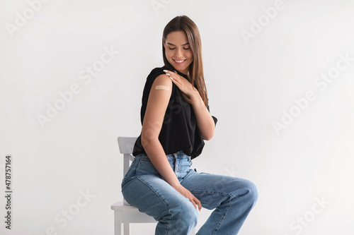 Smiling Caucasian woman showing shoulder with band aid after coronavirus vaccine injection on light studio background