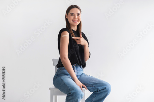Happy young female patient pointing at her arm with band aid after coronavirus vaccine shot on light studio background