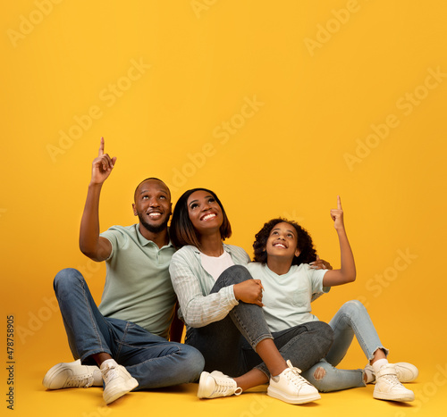 Look there. Happy african american family pointing fingers up at empty space, sitting on floor over yellow background