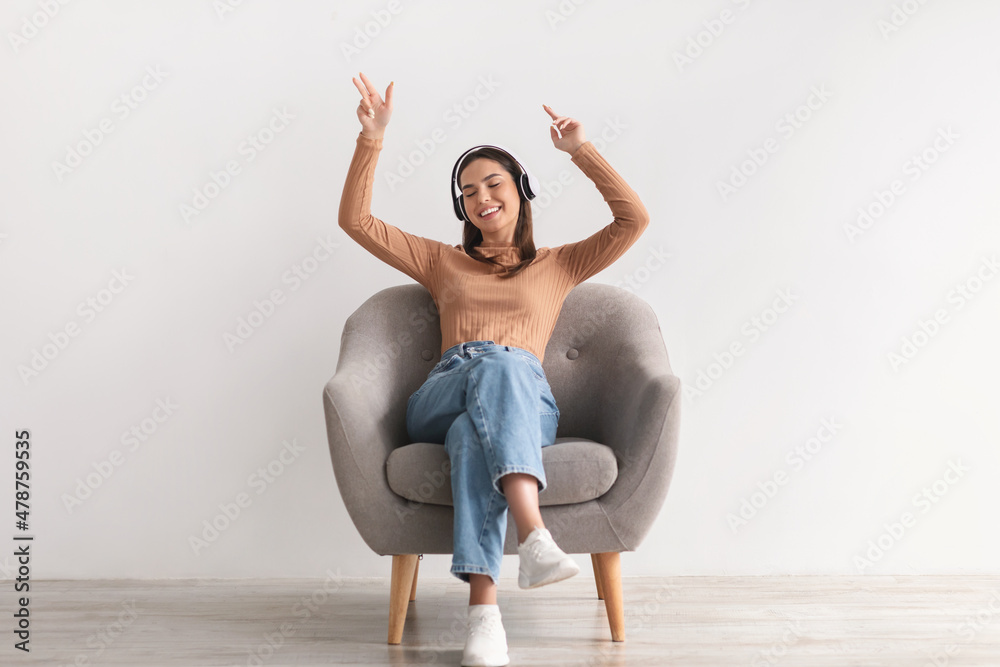 Cool young lady sitting in armchair, listening to music in wireless headphones, dancing to her favorite song