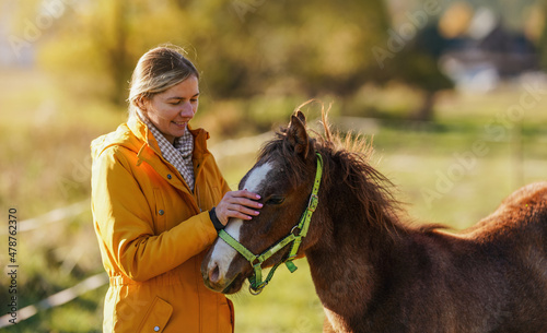 Small brown Arabian horse foal, young woman in yellow orange jacket standing next, blurred sun lit autumn trees background