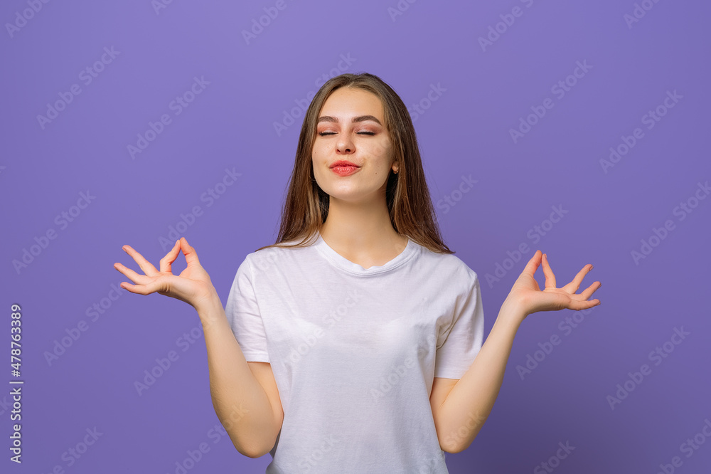 Portrait of calm beautiful girl meditating in white tshirt, smiling while raising hands with zen gestures, her eyes closed, tries to relax after long hours of study, standing against purple background Portrait of calm beautiful girl meditating in white tshirt, smiling while raising hands with zen gestures, her eyes closed, tries to relax after long hours of study, standing against purple background