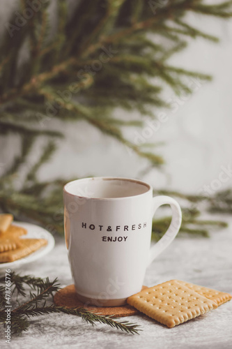 Light ceramic mug framed by green spruce branches, next to a plate with cookies.The inscription on the mug " hot fresh enjoy" 