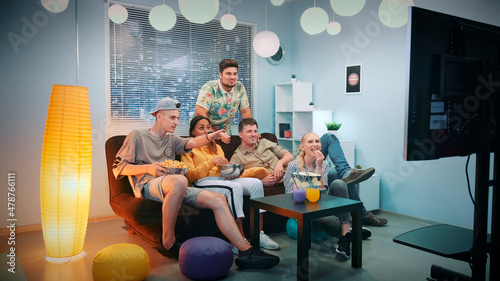 Diverse group of friends watching sports on TV while eating snacks at home. There are skyscrapers in the background. They are disappointed with their team play. Elements of this image furnished by