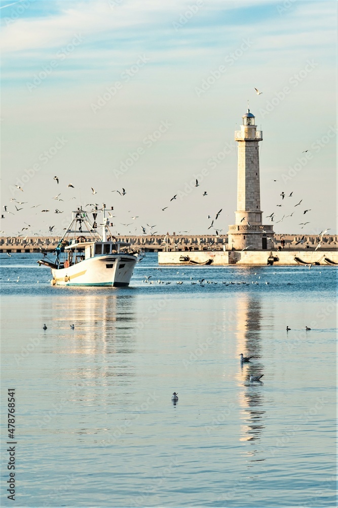 Old fishing boat returning from fishing enters the port at sunset accompanied by a flock of seagulls. With beautiful lighthouse in the background, vertical