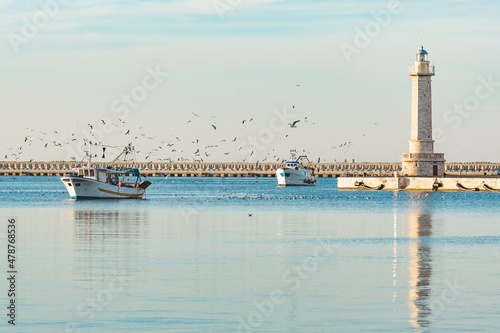 Old fishing boats returning from fishing enters the port at sunset accompanied by a flock of seagulls. With beautiful lighthouse in the background