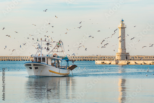 Fotografie Old fishing boat returning from fishing enters the port at sunset accompanied by a flock of seagulls