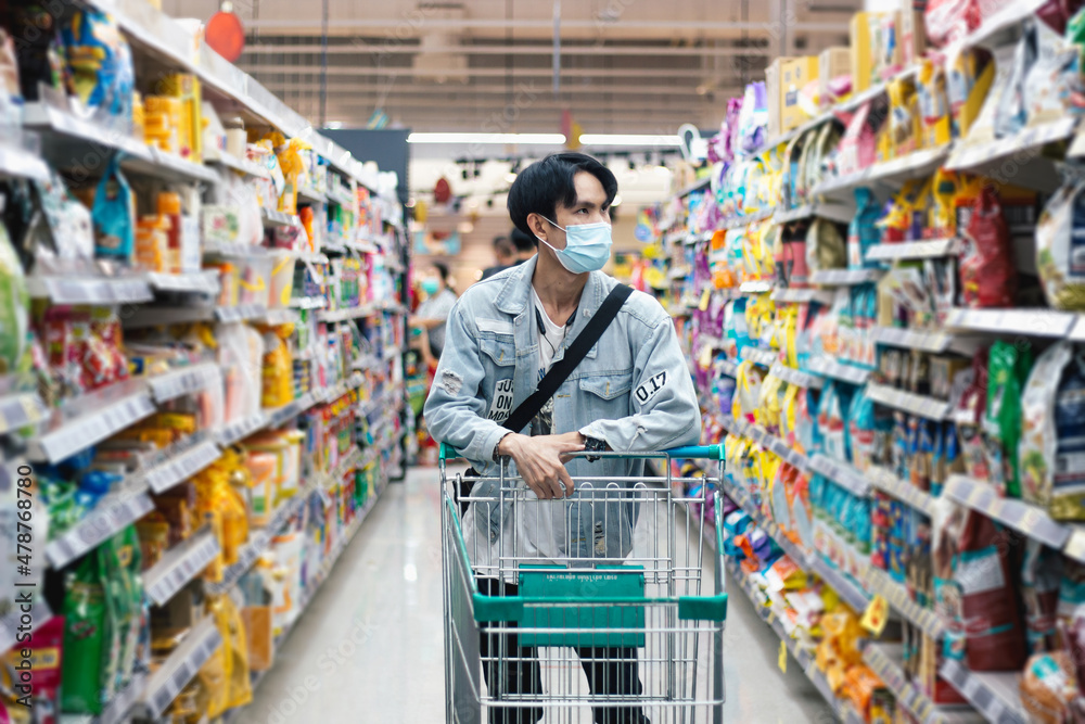 Asian man shopping for groceries walks with a shopping cart wearing a protective mask in a modern supermarket. Male customers who shop in indoor food stores. spot focus - obrazy, fototapety, plakaty 