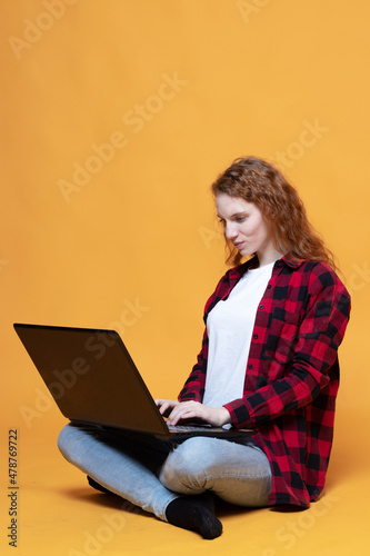 red-haired girl in a plaid shirt sits with a laptop on an orange background