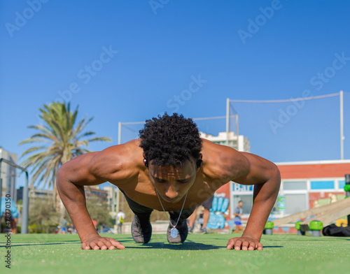 A shirtless dark skin tone guy doing push ups at the morning on a excersise public area.