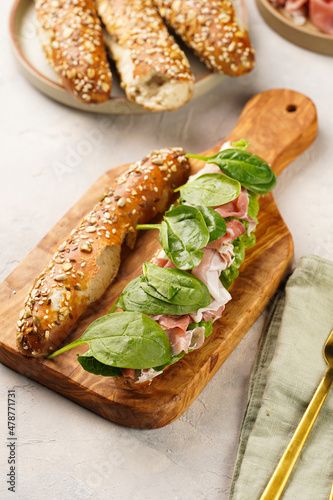 Long sandwich in brown lye bread sticks garnished with oats , pork slices prosciutto, hard cheese gouda and fresh greens on wooden board, beige surface