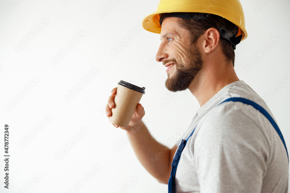 Side view portrait of smiling handyman in uniform and hard hat drinking coffee during a break while standing indoors Side view portrait of smiling handyman in uniform and hard hat drinking coffee during a break while standing indoors