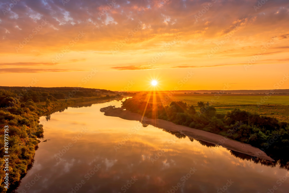 Fototapeta premium Scenic view at beautiful sunset or sunrise on a shiny river with green bushes on sides, golden sun rays, calm water ,deep blue cloudy sky and forest on a background, spring landscape