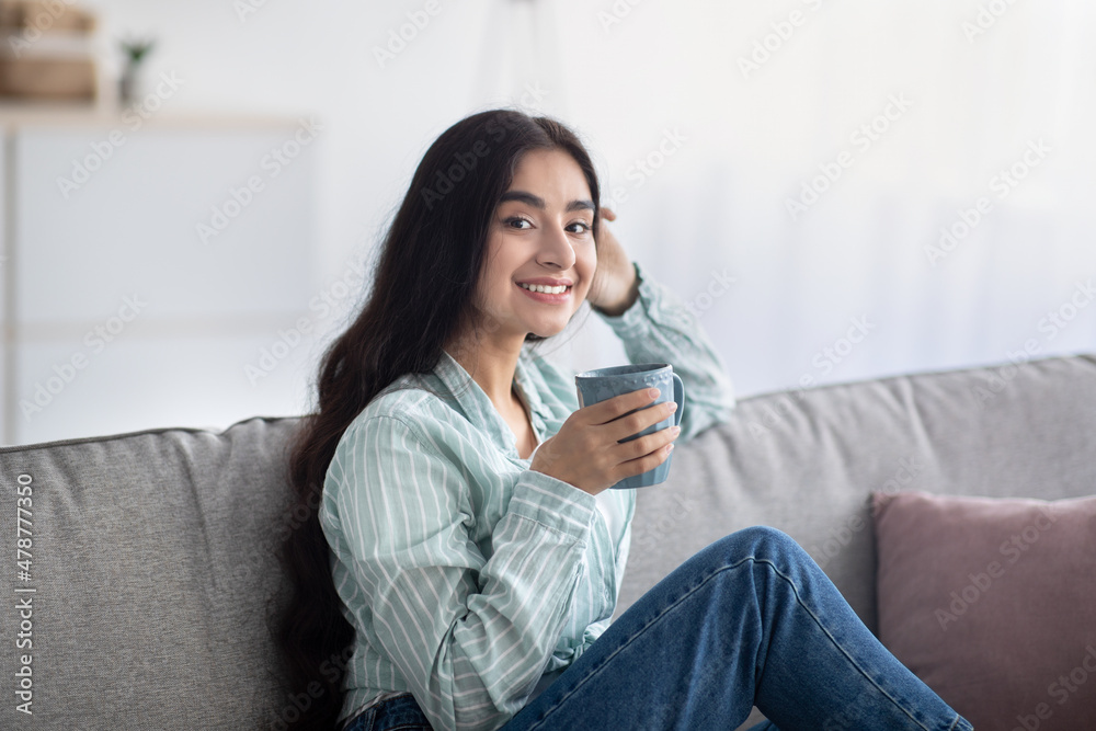 Portrait of young Indian woman having coffee break, sitting on sofa with cup of hot beverage at home