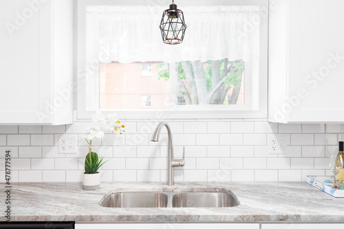 Kitchen sink detail shot with a marble countertop, white cabinets, stainless steel faucet and sink, and a light hanging in front of the window.	 photo