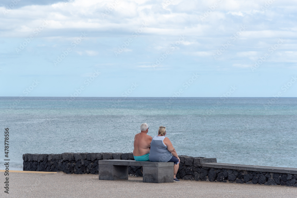Una pareja de personas mayores sentadas en un banco mirando el mar en una playa de Lanzarote.