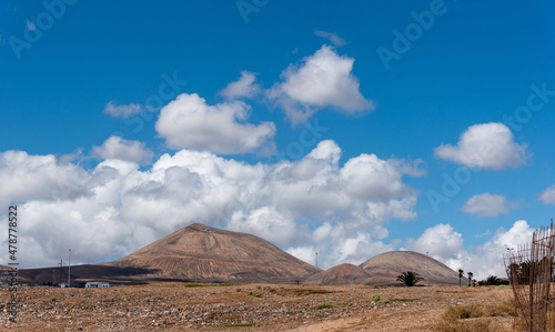 Paisaje volcanico con montañas al fondo y terreno arido con nubes blancas y cielo azul en Lanzarote, Islas Canarias.