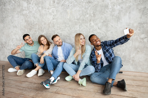 Full length of millennial students taking selfie with smartphone, sitting near grey studio wall, copy space