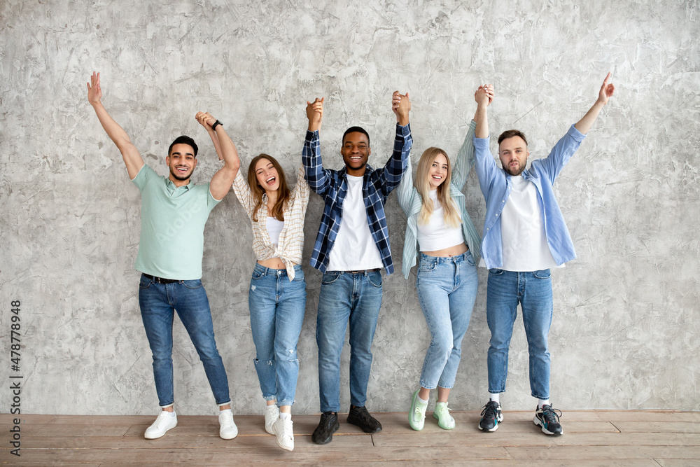 Obraz premium Full length of diverse friends holding hands up, feeling happy, looking at camera against grey studio wall