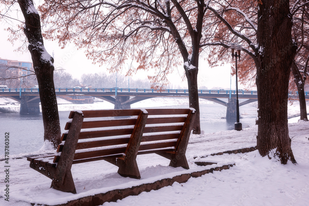 Winter cityscape, famous linden alley on the embankment of Uzh river with a snow, wooden bench, linden trees and road bridge on background, Uzhhorod, Ukraine
