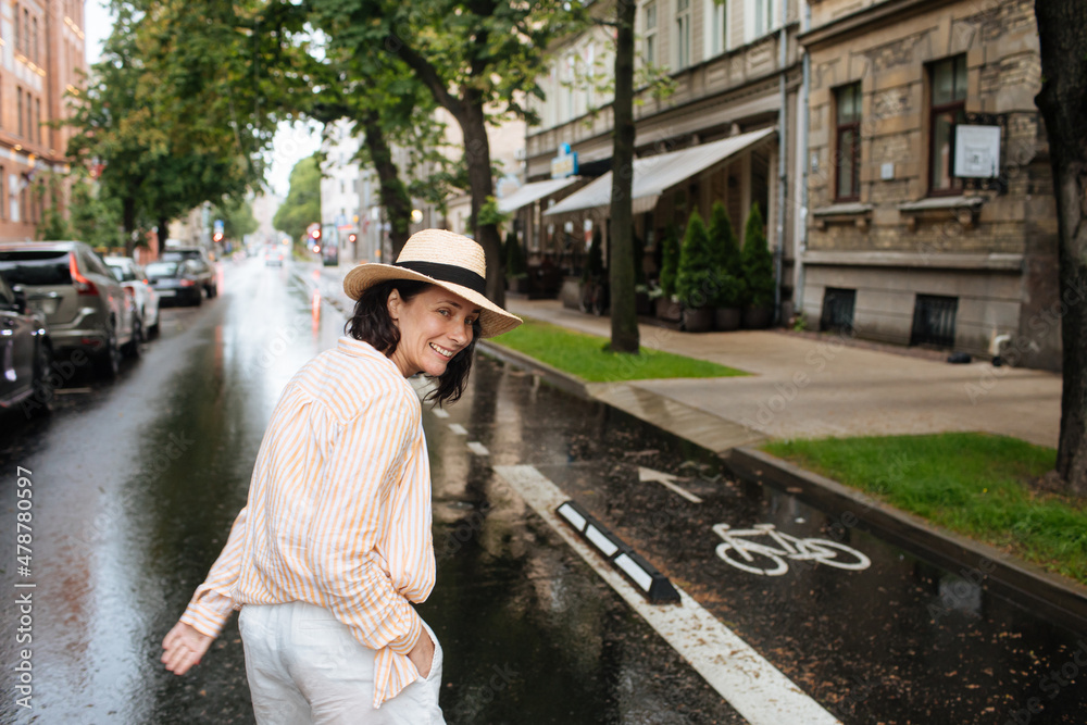 Portrait of Woman walking in the middle of the street. Female unsing a mobile phone in summer rain. City lifestyle fashion. Woman holding cell phone and wearing summer clothes and hat.