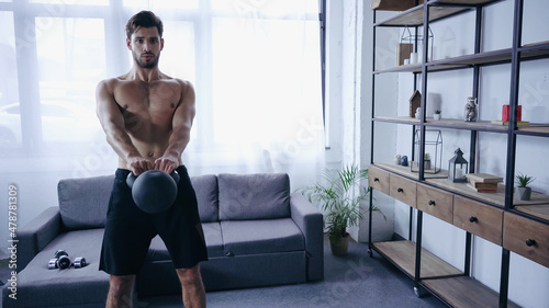 shirtless sportsman training with kettlebell near sofa with dumbbells at home
