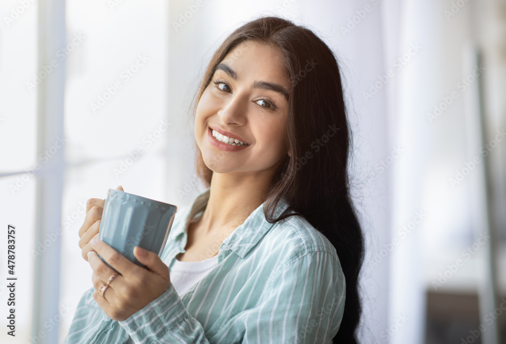 Portrait of pretty Indian lady standing near window with cup of hot drink, smiling at camera, indoors Portrait of pretty Indian lady standing near window with cup of hot drink, smiling at camera, indoors