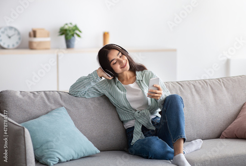 Joyful Indian lady in headphones sitting on couch and listening to music on smartphone at home, full length