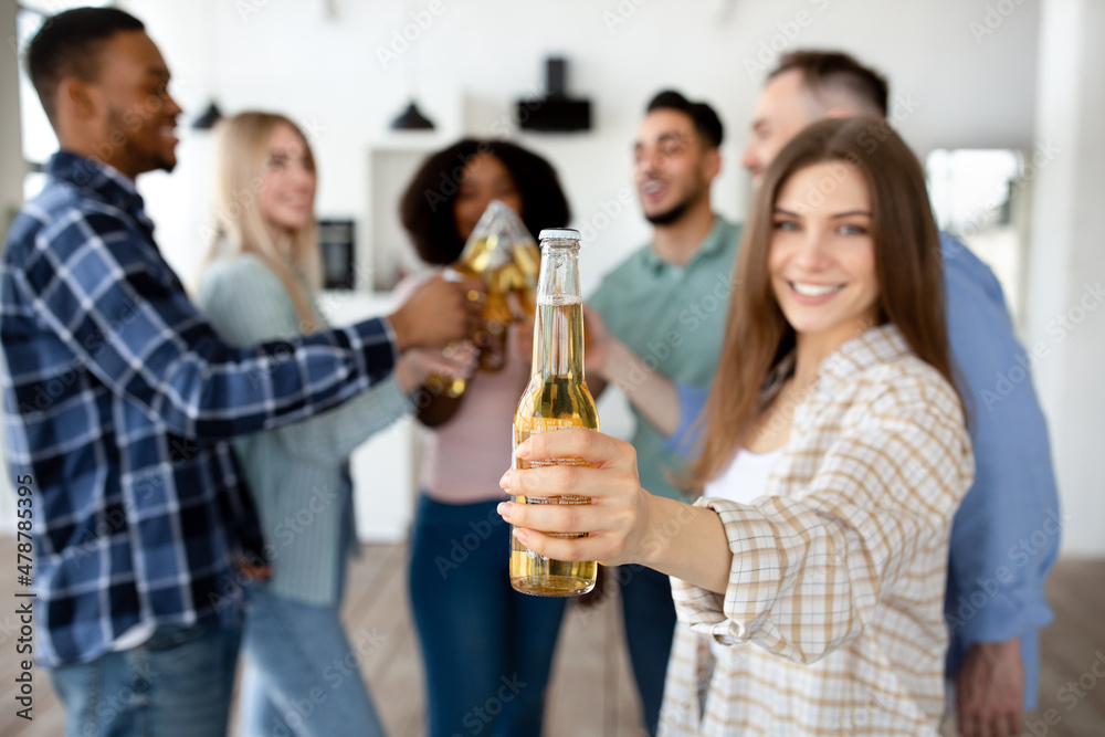 Young Caucasian woman with her multiracial friends toasting with beer bottles, celebrating something at home, closeup