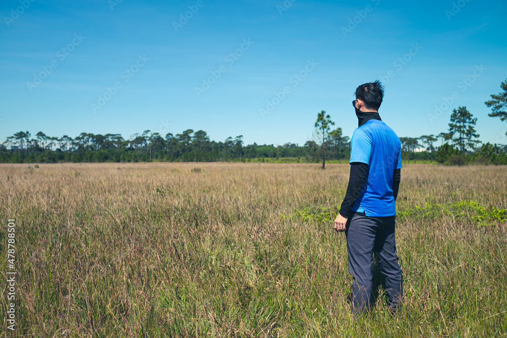 Young man standing in a deserted meadow He was looking out into the vast grassland. with a background of sky and pine trees., Rear view , man portrait on landscape , National Park ,Thailand