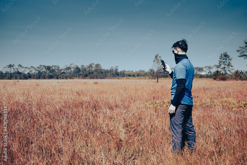 A young man wearing sunglasses and a windproof mask stands in a barren meadow. He was checking the weather forecast on his cell phone , side view , man portrait on landscape , National Park ,Thailand