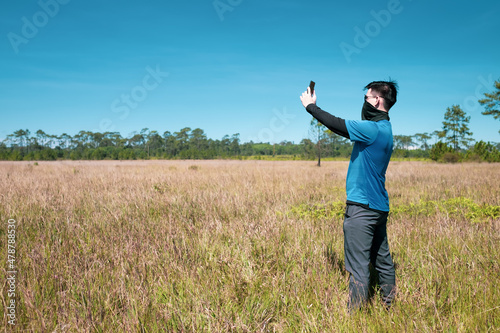 A young man wearing sunglasses and a windproof mask in hand smartphone selfie stands in the meadow , side view , man portrait on landscape , National Park ,Thailand