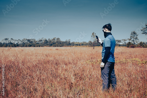 A young man wearing sunglasses and a windproof mask stands in a barren meadow. He was checking the weather forecast on his cell phone , side view , man portrait on landscape , National Park ,Thailand © AmazingSingsong