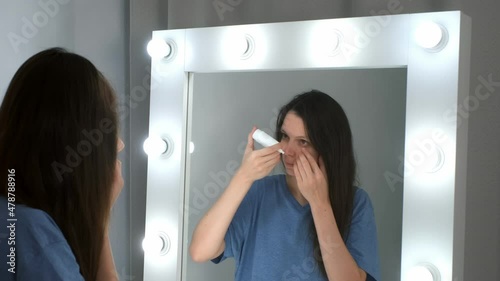 Woman is dripping drops in her eyes in front of the mirror at home. She is looking at her reflection in mirror. Eye fatigue, manager syndrome, exhaustion, stress, overwork, sleep disorder concepts. photo
