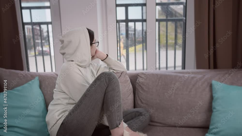 Girl in the hood looks thoughtfully out the window while sitting on the sofa Girl in the hood looks thoughtfully out the window while sitting on the sofa